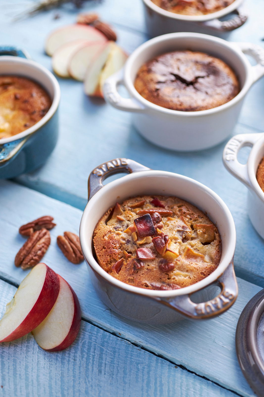 Vogelperspektive auf ein Set von Mini-Cocottes in verschiedenen Farben, gefüllt mit gebackenem Dessert, umgeben von Apfelscheiben und Pekannüssen auf einem blauen Holztisch.