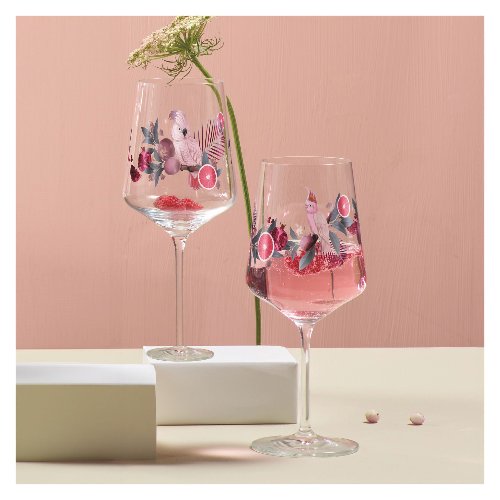 Zwei Weingläser mit tropischem Design, verziert mit Papageien und exotischen Früchten, auf einem Tisch mit rosa Hintergrund. Seitliche Perspektive.