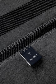 Detailaufnahme aus schräger Draufsicht: graue JOOP! Wohndecke mit eingewebtem Muster, eingefasster Kante und sichtbarem Markenlabel