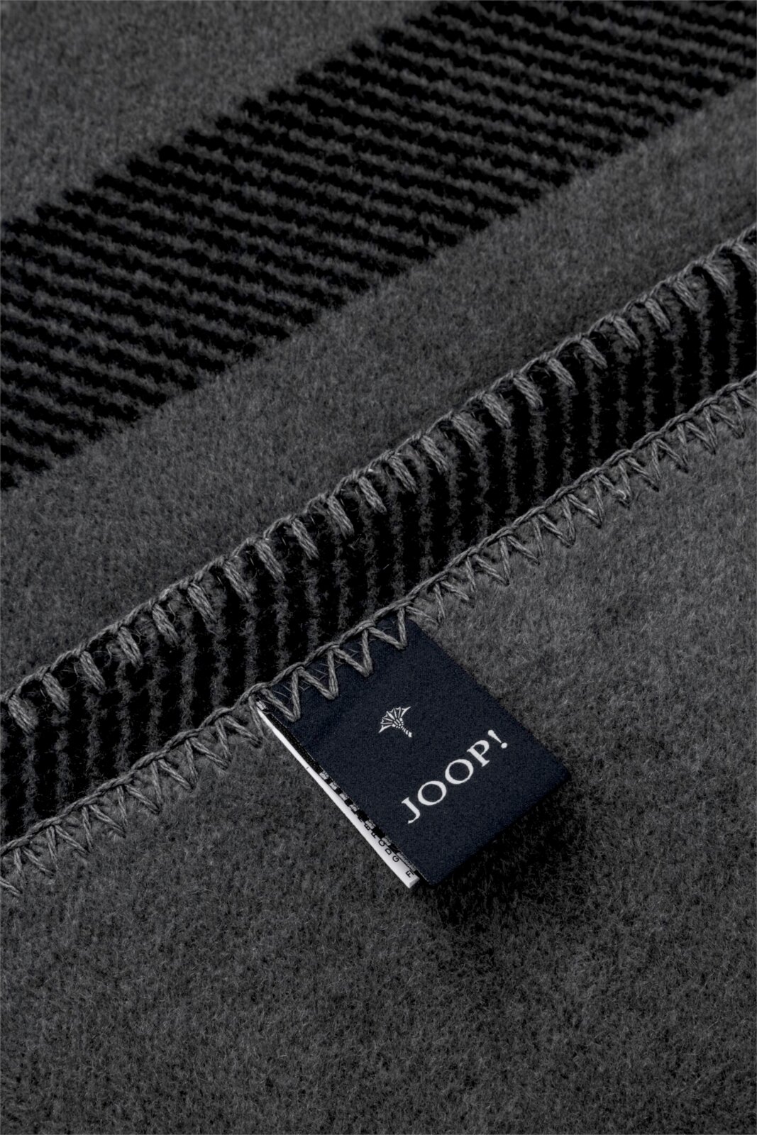 JOOP! Wohndecke 150 x 200cm SHUTTER Detailaufnahme aus schräger Draufsicht: graue JOOP! Wohndecke mit eingewebtem Muster, eingefasster Kante und sichtbarem Markenlabel