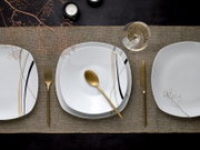 Draufsicht auf ein modern gedecktes Tisch-Setting mit weißem, quadratisch abgerundetem Porzellanteller mit schwarzen und goldenen Linien, goldenem Besteck, Glas und Kerze auf Leinenläufer.