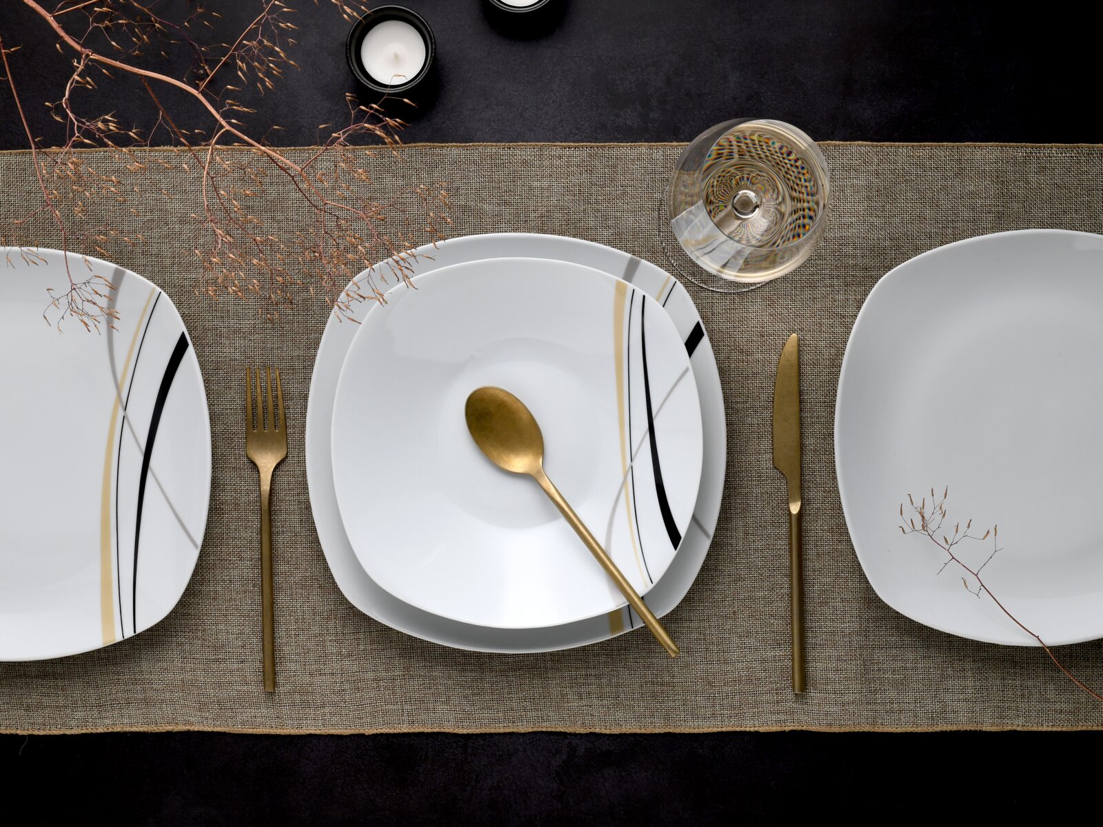 Draufsicht auf ein modern gedecktes Tisch-Setting mit weißem, quadratisch abgerundetem Porzellanteller mit schwarzen und goldenen Linien, goldenem Besteck, Glas und Kerze auf Leinenläufer.