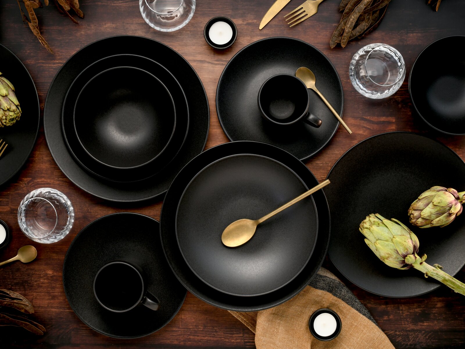 Schwarzes, mattes Geschirrset mit Tellern, Schalen und Tassen, stilvoll arrangiert mit goldenem Besteck, Gläsern und Artischocken auf einem dunklen Holztisch, aus der Vogelperspektive