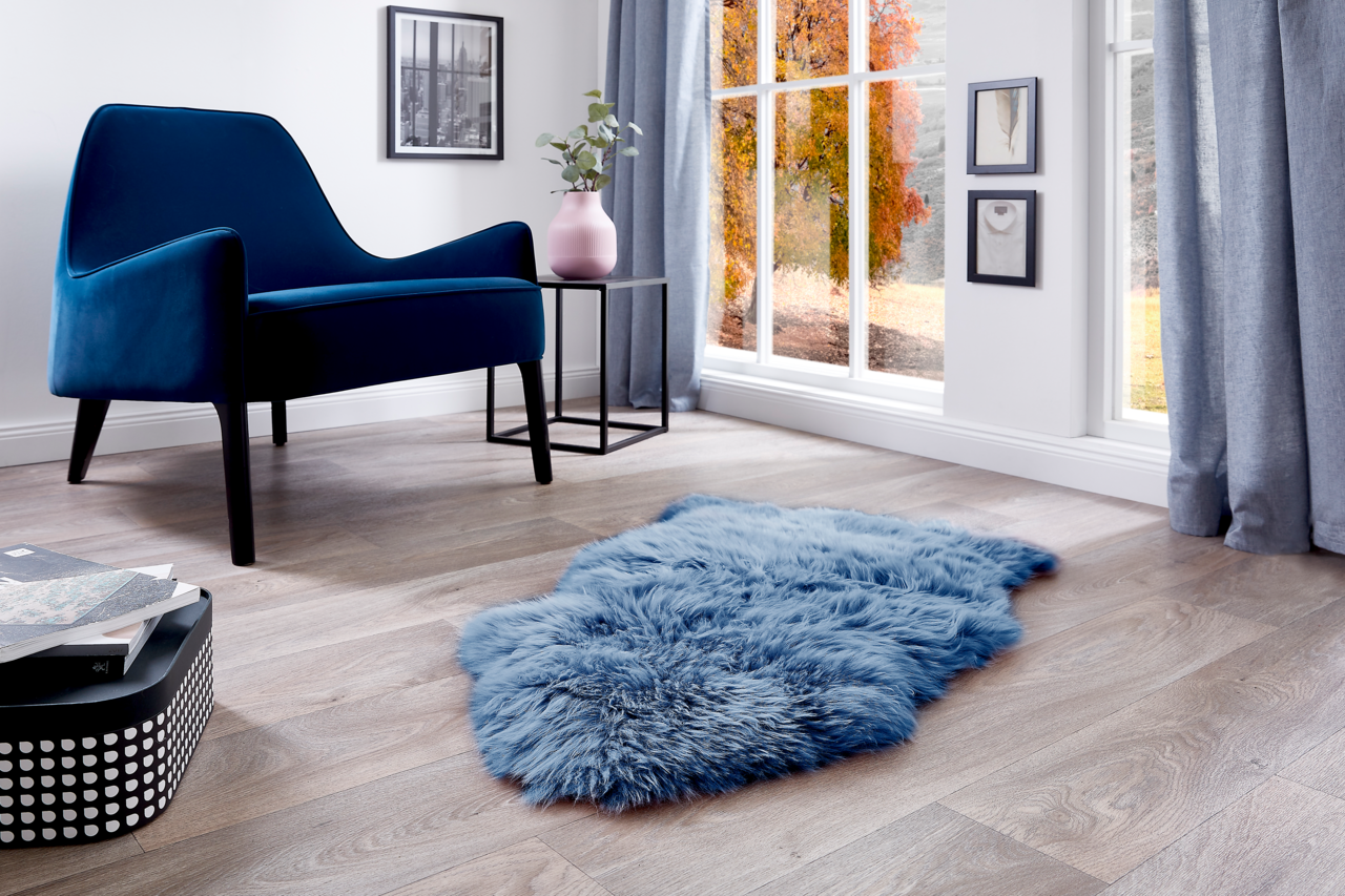 Blaues Schaffell auf einem Holzboden in einem modernen Wohnzimmer mit einem blauen Sessel und einem Fenster mit herbstlicher Aussicht, aus der Vogelperspektive.