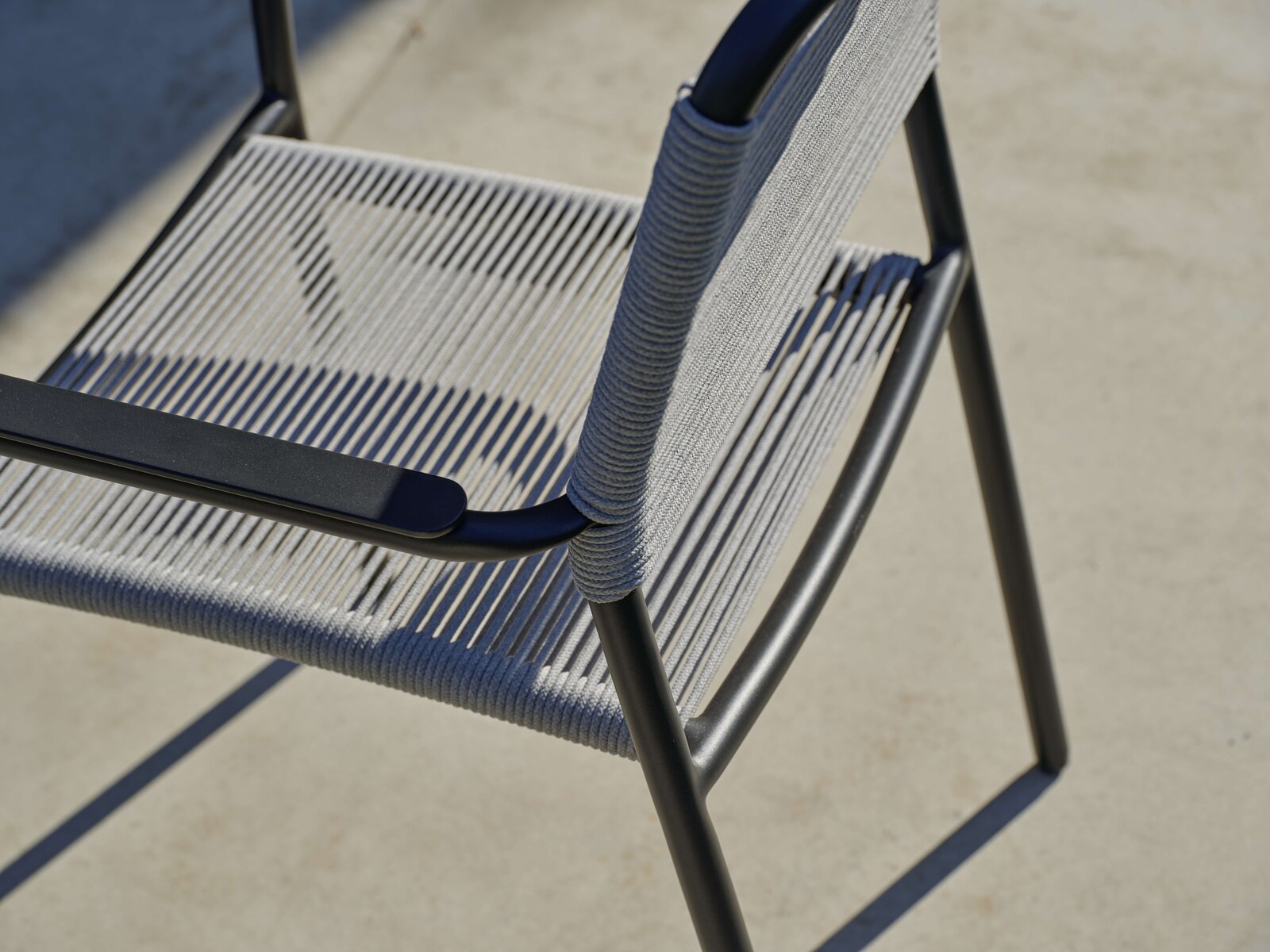 Nahaufnahme eines modernen Stapelsessels mit schwarzem Metallrahmen und grauer Seilbespannung, von oben betrachtet.