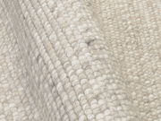 Nahaufnahme eines handgewebten Teppichs mit grober Struktur in neutralen Farbtönen, seitliche Perspektive.