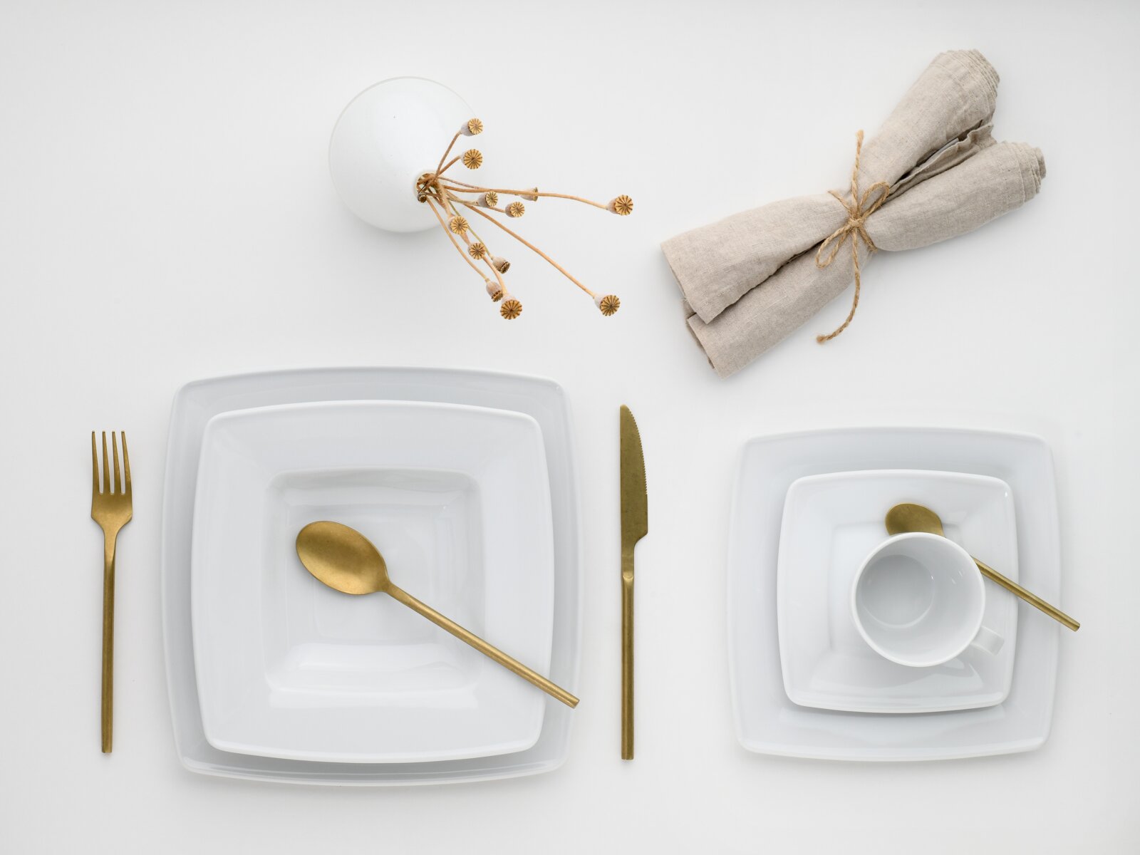 Minimalistisches Tischgedeck mit weißen quadratischen Tellern, goldfarbenem Besteck, weißer Tasse und beigefarbenen Stoffservietten mit Trockenblumen auf weißem Hintergrund, Draufsicht von oben