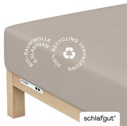 Spannbettlaken aus Bio-Baumwolle und Elasthan in Beige, auf einer Matratze von schräg oben fotografiert, mit Hinweis auf 100% Recycling-Verpackung.