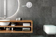 Modernes Badezimmer mit rundem, hinterleuchtetem Spiegel, Waschtisch mit Holzregal und wandhängendem WC vor grauen Fliesen, Frontansicht mit leicht seitlicher Perspektive.