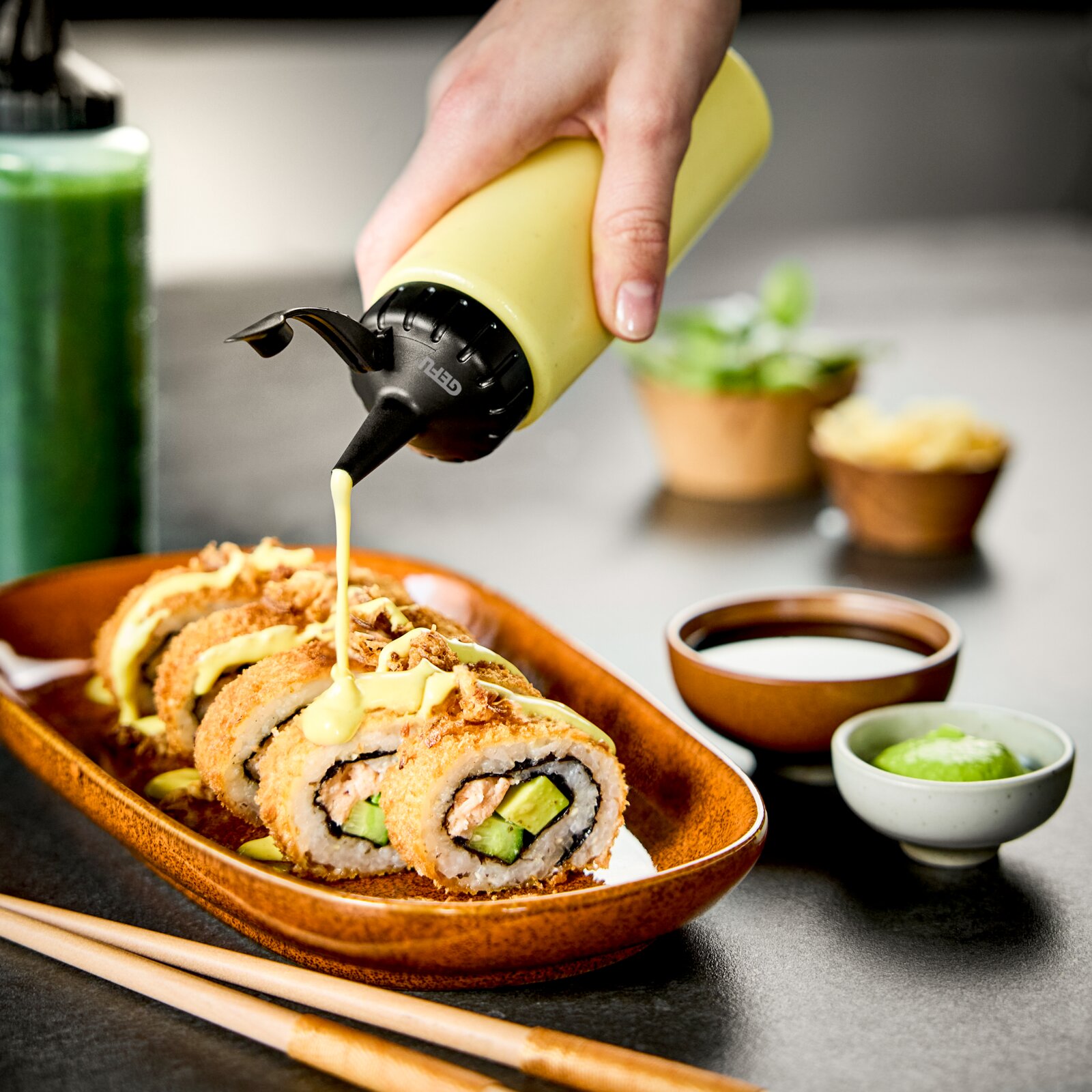 Hand drückt Sauce aus einer gelben Dosierflasche auf Sushi-Rollen, seitliche Perspektive.