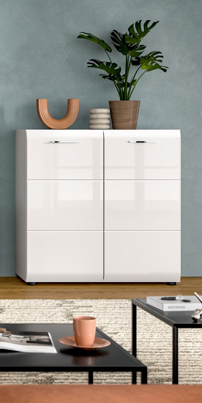 Frontale Ansicht eines weißen, hochglänzenden Sideboards mit sechs Klappen und silbernen Griffen vor grauer Wand, dekoriert mit Pflanze und Vasen; im Vordergrund Couchtische mit Tasse und Zeitschriften.