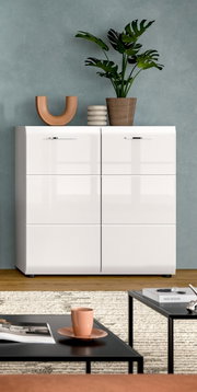 Frontale Ansicht eines weißen, hochglänzenden Sideboards mit sechs Klappen und silbernen Griffen vor grauer Wand, dekoriert mit Pflanze und Vasen; im Vordergrund Couchtische mit Tasse und Zeitschriften.
