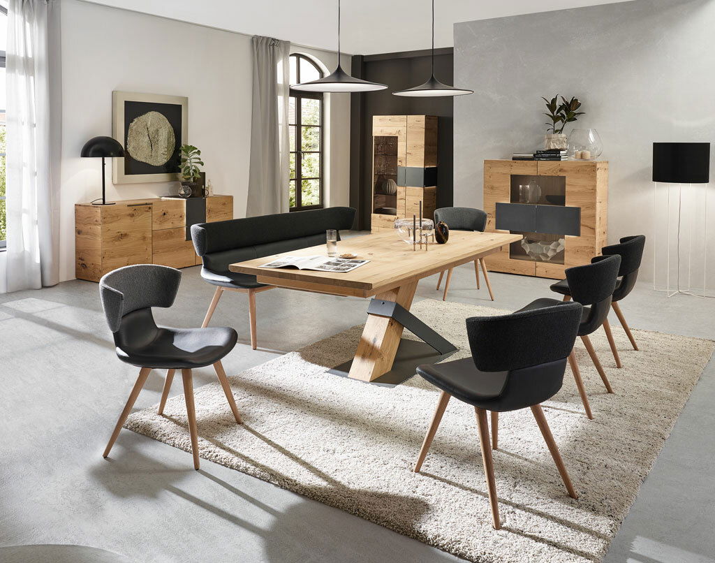 Moderne Esszimmermöbel in einem hellen Raum, bestehend aus einem großen Holztisch mit schwarzem Metallgestell, umgeben von schwarzen Stühlen und einer Bank, mit einem Sideboard und einer Vitrine im Hintergrund. Perspektive: Frontalansicht des Essbereichs.