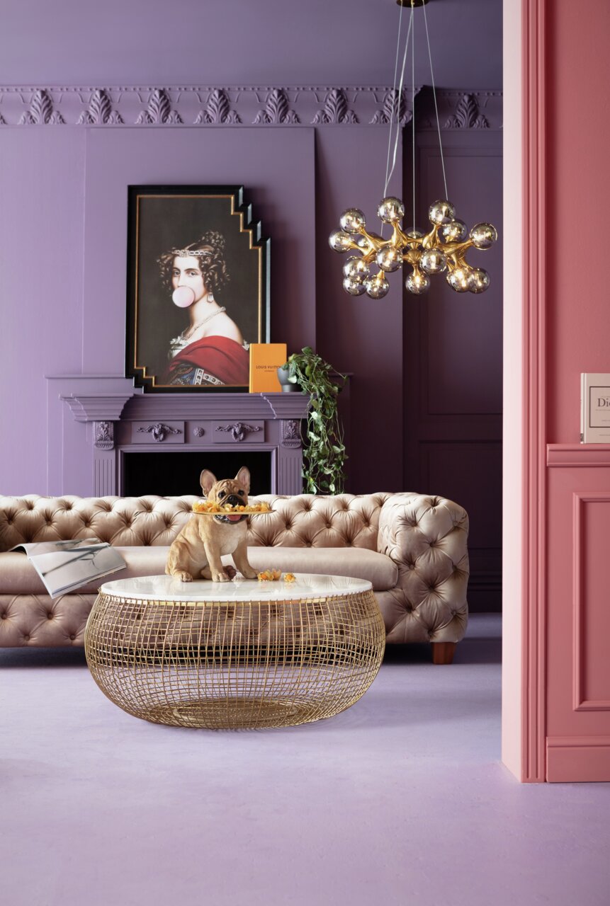 Deko-Figur eines sitzenden French Bulldog Butlers mit Tablett im Maul, auf einem runden Tisch in einem stilvoll eingerichteten Raum mit lila Wänden und einem eleganten Sofa. Perspektive: Frontalansicht.
