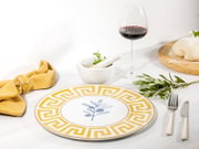 Weißer Speiseteller mit goldfarbenem Mäander-Rand und blauem Olivenzweig-Motiv, aufgedeckt auf weißem Tisch mit Besteck, Kräutern und Rotweinglas, fotografiert in leicht schräger Draufsicht.