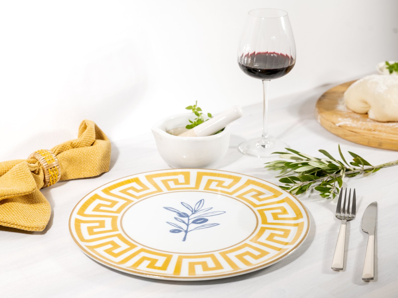 Weißer Speiseteller mit goldfarbenem Mäander-Rand und blauem Olivenzweig-Motiv, aufgedeckt auf weißem Tisch mit Besteck, Kräutern und Rotweinglas, fotografiert in leicht schräger Draufsicht.