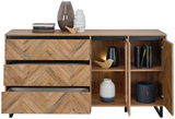 Sideboard aus Holz mit Fischgrätmuster, drei Schubladen und zwei offenen Türen, dekoriert mit Vasen, Büchern und einer Lampe, Frontalansicht.