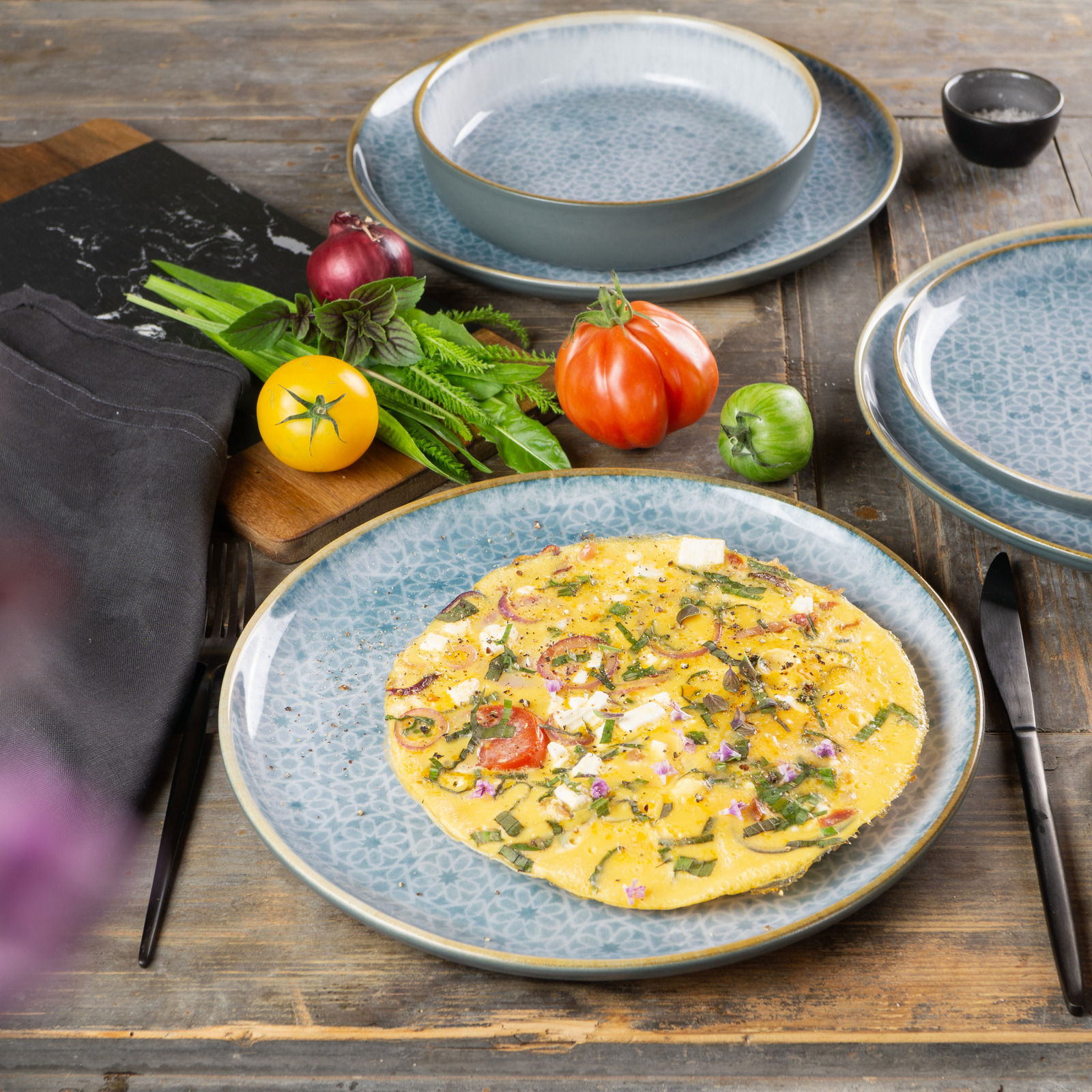 Geschirrset Terra 18-tlg. Azur mit einem Teller im Vordergrund, darauf ein Omelett, umgeben von frischen Zutaten wie Tomaten und Kräutern, auf einem rustikalen Holztisch arrangiert.