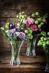 Drei elegante Kristallvasen mit bunten Blumensträußen, auf einem rustikalen Holztisch, aus leicht erhöhter Perspektive fotografiert.