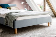 Een gestoffeerd bed van lichtblauwe stof met houten poten, gezien vanaf de zijkant, versierd met kussens en een deken in een moderne slaapkamer.