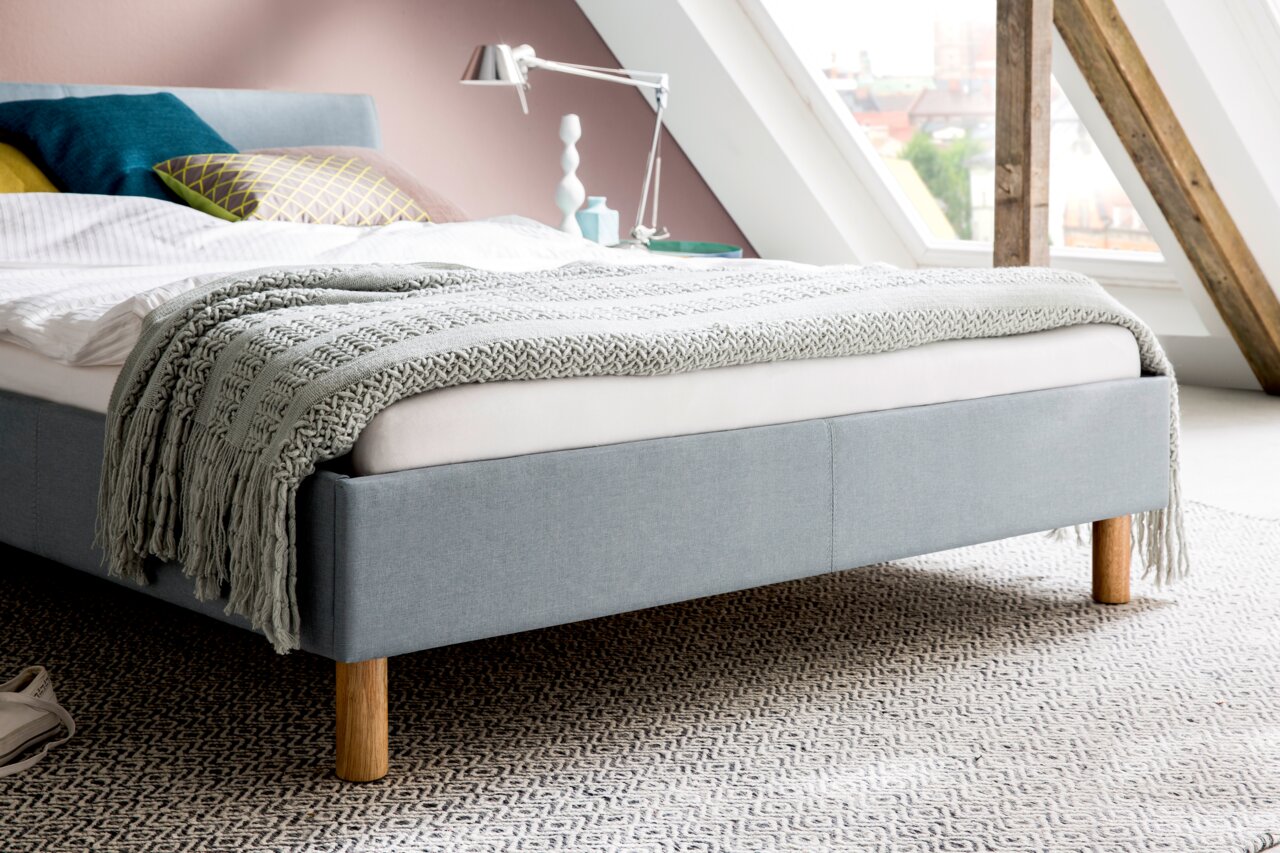 Een gestoffeerd bed van lichtblauwe stof met houten poten, gezien vanaf de zijkant, versierd met kussens en een deken in een moderne slaapkamer.