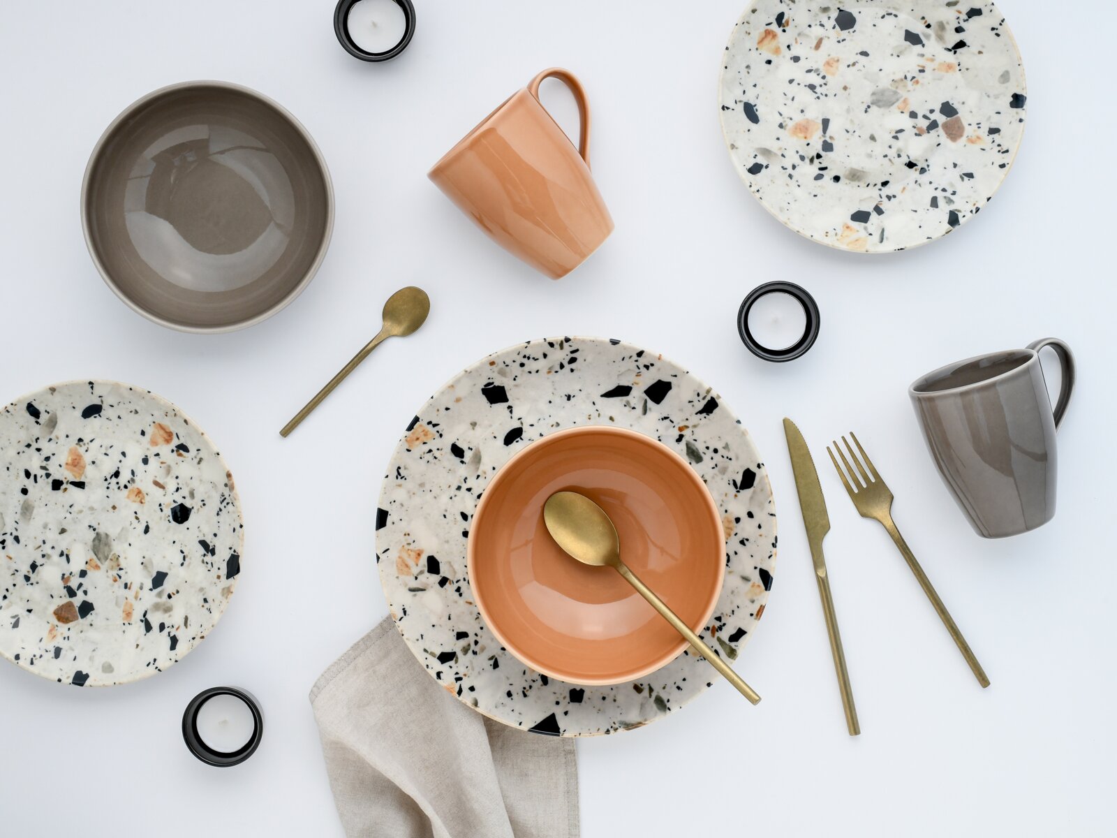 Flatlay aus der Vogelperspektive: modernes Geschirr-Set mit gesprenkelten Terrazzo-Tellern, grauer Schale, orangefarbener Schüssel und Tasse, grauer Tasse sowie goldfarbenem Besteck und kleinen Teelichtern auf hellem Hintergrund