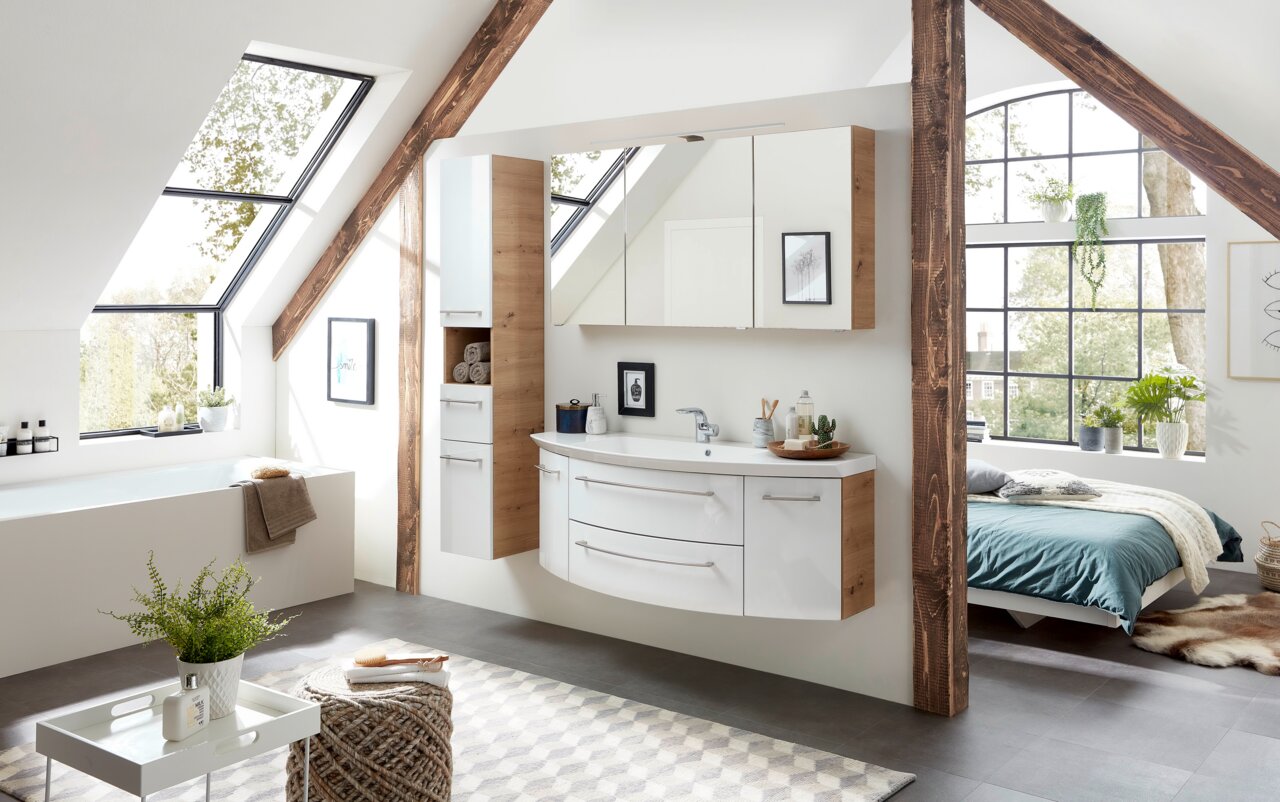 Sadena Badkombination 3-tlg. PLAKA Moderne Badkombination in einem hellen Dachgeschossbad mit Holz- und Weißtönen, bestehend aus einem Waschtisch mit Schubladen und einem Spiegelschrank, seitliche Perspektive.