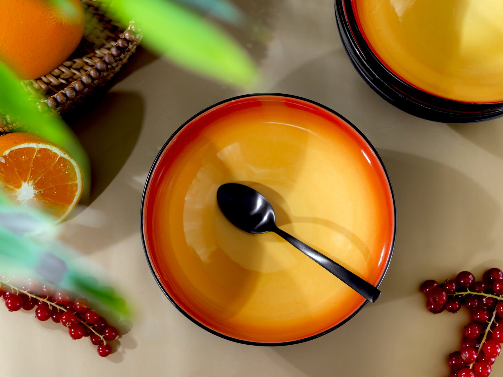 Orangefarbene Schale mit schwarzem Rand und schwarzem Löffel, aus der Vogelperspektive zwischen Orangen und roten Johannisbeeren arrangiert