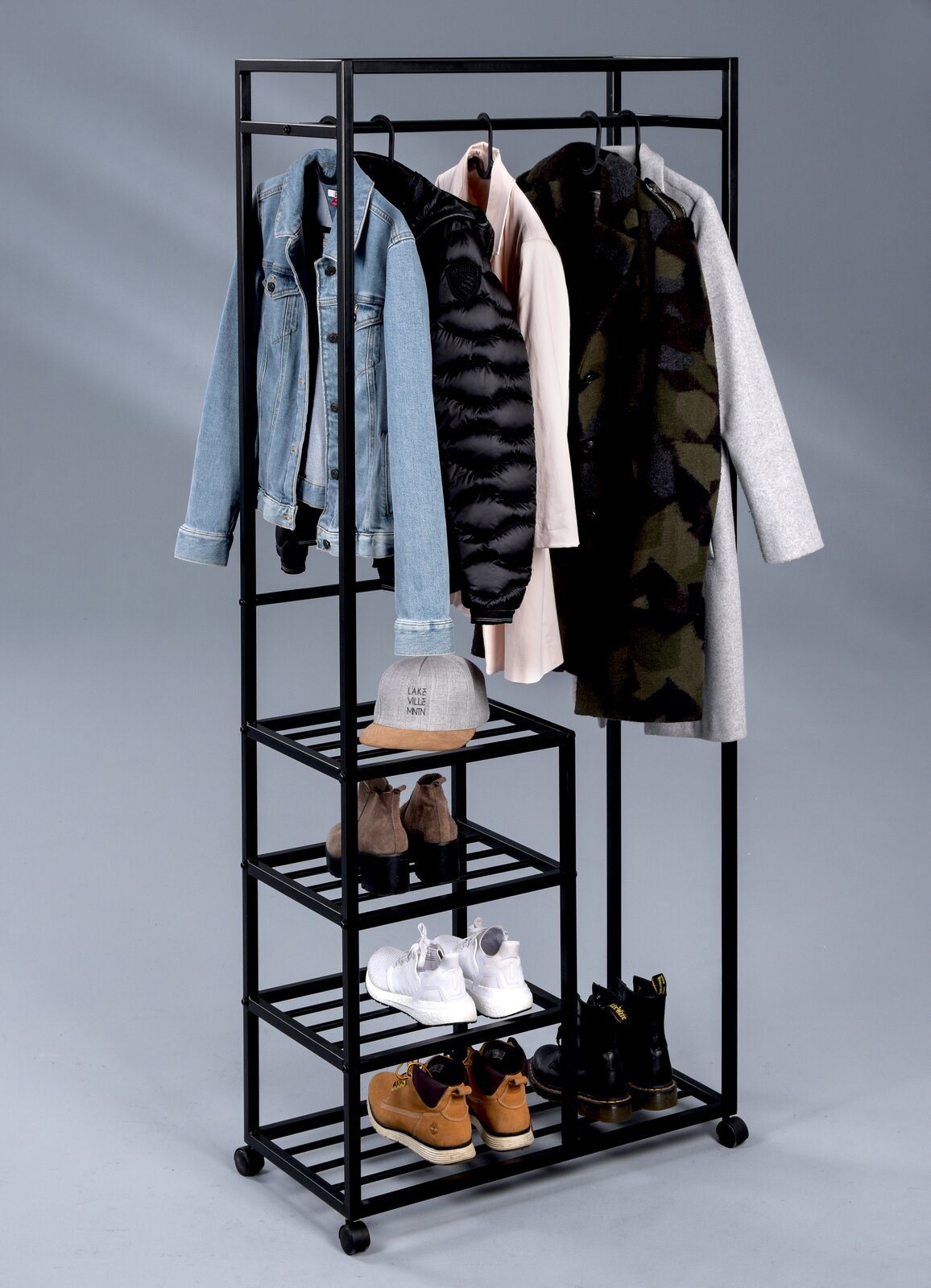 Garderobenwagen aus schwarzem Metall mit mehreren Regalböden und Kleiderstange, darauf hängen Jacken und Mäntel; Schuhe und eine Mütze sind auf den Regalen platziert. Perspektive: Frontalansicht.