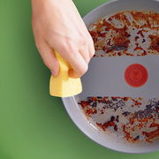 Nahaufnahme einer Hand, die mit einem gelben Schwamm eine verschmutzte Wokpfanne reinigt. Die Pfanne zeigt deutliche Gebrauchsspuren und ist von oben zu sehen.