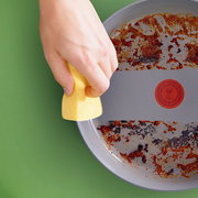 Nahaufnahme einer Hand, die mit einem gelben Schwamm eine verschmutzte Wokpfanne reinigt. Die Pfanne zeigt deutliche Gebrauchsspuren und ist von oben zu sehen.