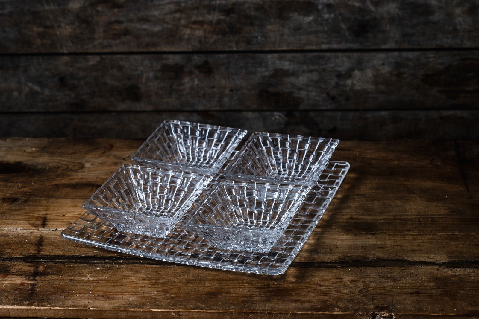 Perspektive von oben auf ein 5-teiliges Servier-Set Bossa Nova mit vier quadratischen Schalen und einem großen quadratischen Teller, aus klarem Glas mit geflochtenem Muster, auf einem rustikalen Holztisch.