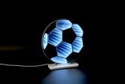 Leuchtende LED-3D-Lampe in Form eines Fußballs mit blauem Infinity-Spiegel-Effekt auf weißem Sockel, schräg von vorn auf schwarzem Hintergrund fotografiert