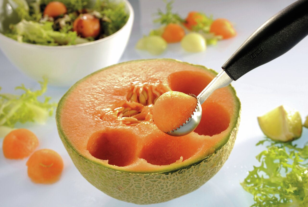Kugelausstecher BOCELI in Nahaufnahme, der eine Melonenkugel aus einer halbierten Melone formt, mit Salat im Hintergrund.