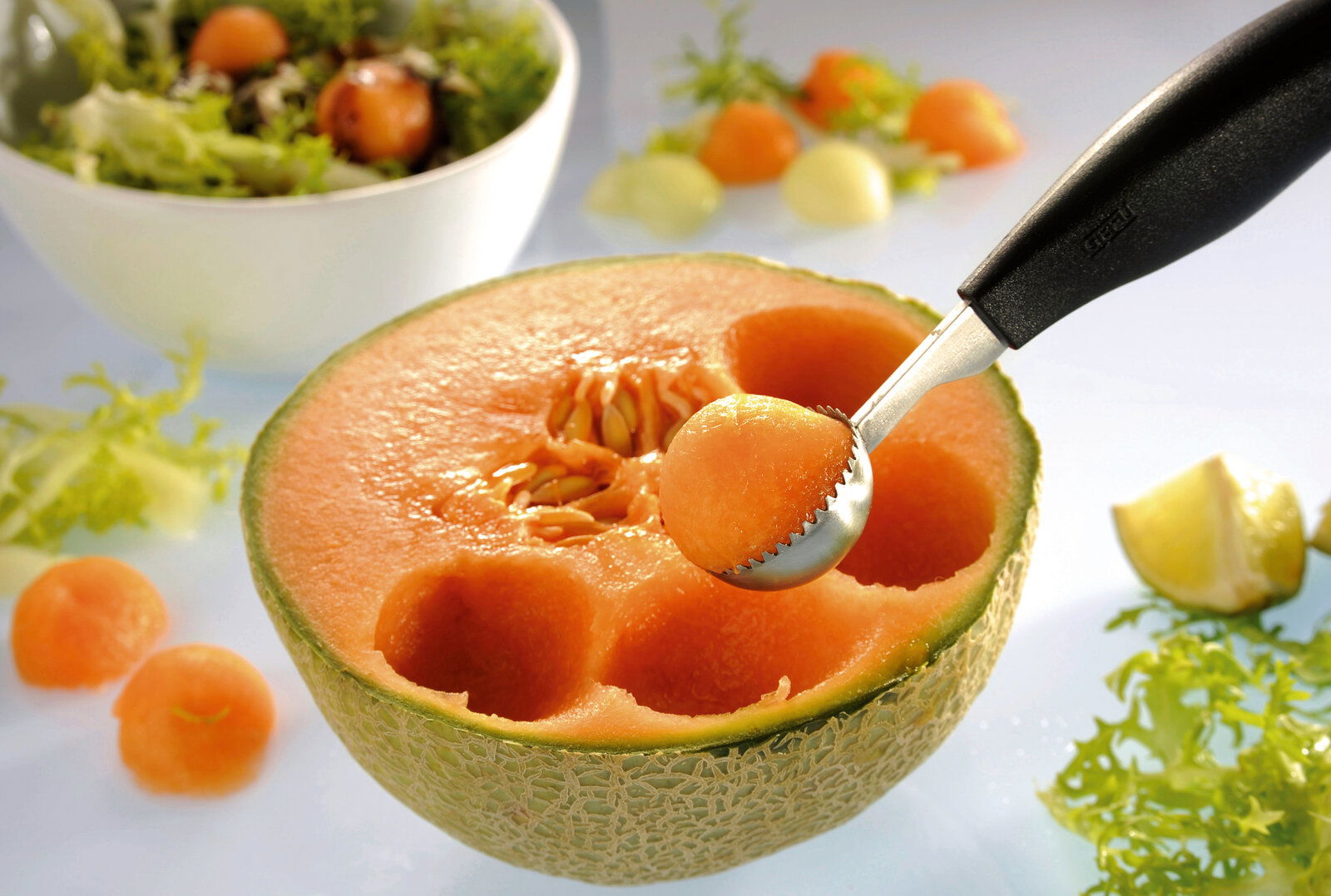 Kugelausstecher BOCELI in Nahaufnahme, der eine Melonenkugel aus einer halbierten Melone formt, mit Salat im Hintergrund.