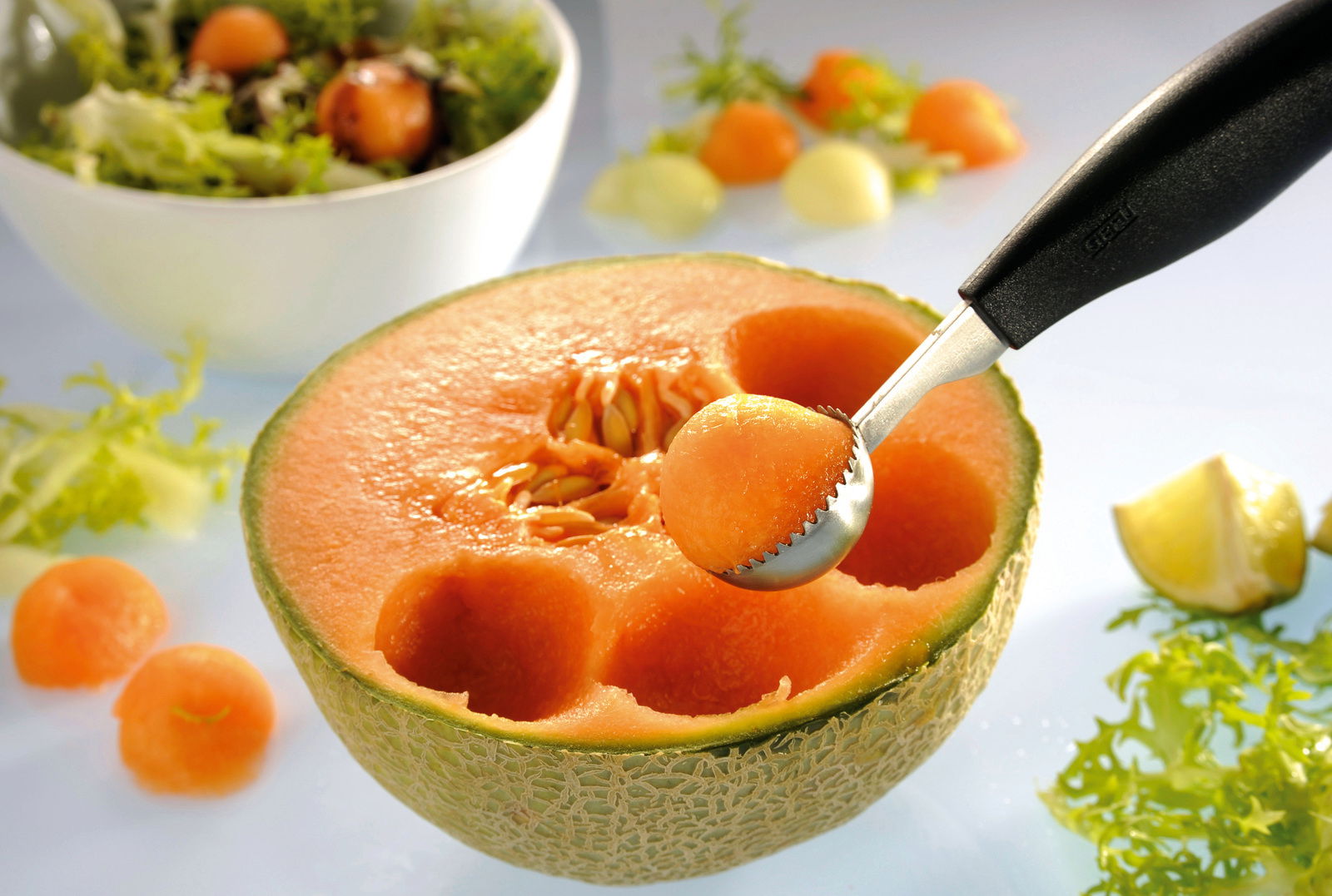 Kugelausstecher BOCELI in Nahaufnahme, der eine Melonenkugel aus einer halbierten Melone formt, mit Salat im Hintergrund.