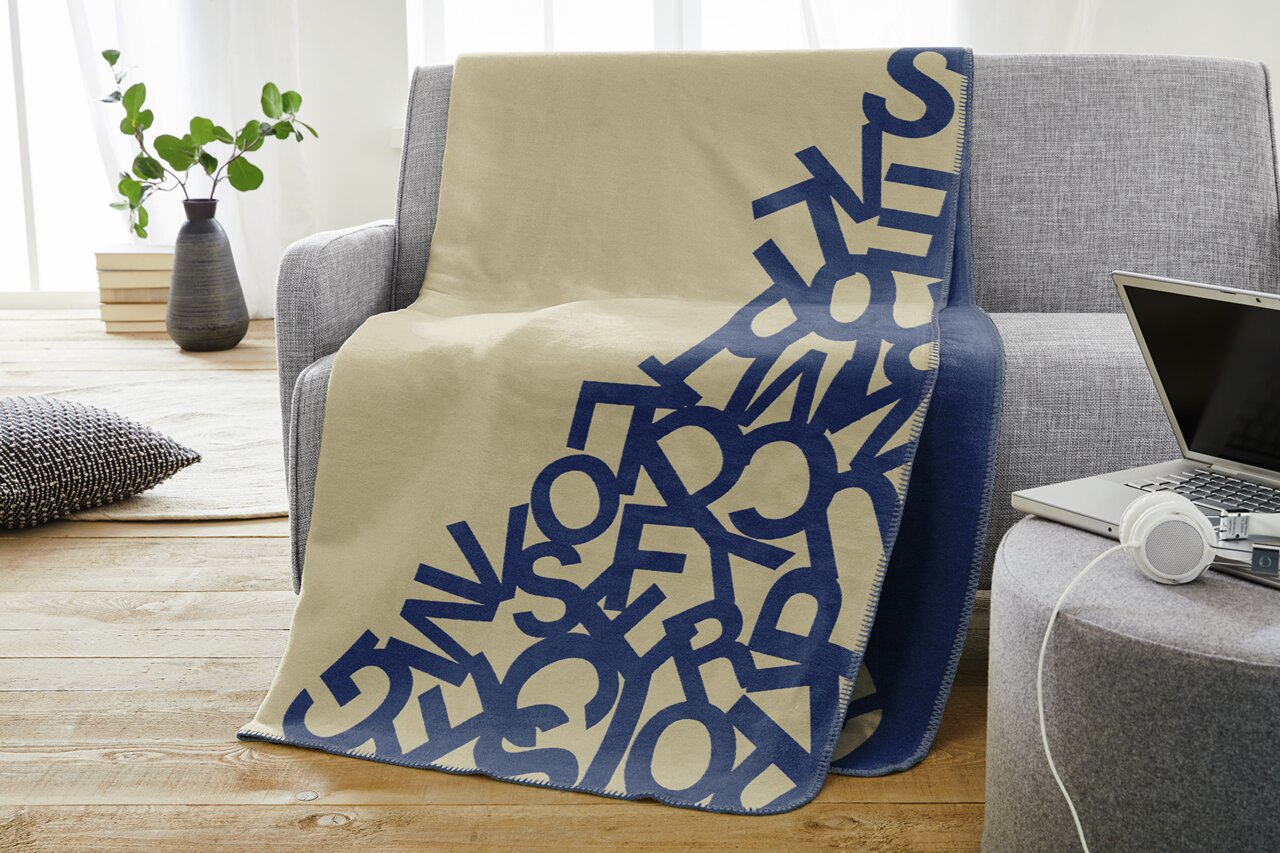 s.Oliver Woonkleed 150x200 cm beige/blauw Jacquarddeken in beige met blauwe letters, gedrapeerd over een grijze bank in een moderne woonkamer.