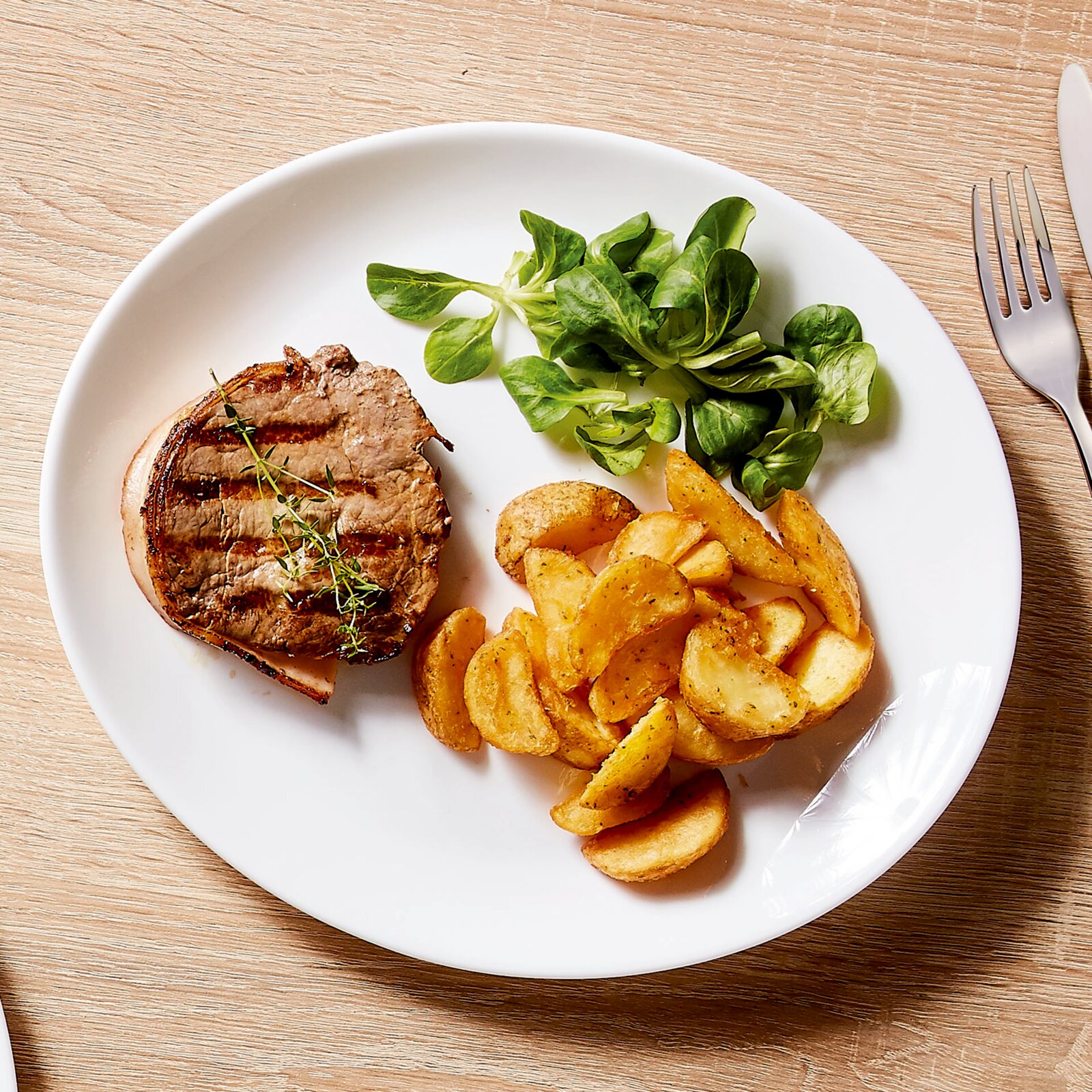 Draufsicht auf einen weißen Teller mit gegrilltem Steak mit Kräutern, goldenen Kartoffelspalten und frischem Feldsalat auf einem Holztisch; rechts liegt eine Gabel.