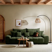 Großes, grünes Sofa mit mehreren Kissen, ein Hund sitzt darauf. Im Vordergrund ein runder Holztisch mit Dekoration. Perspektive: Frontalansicht.