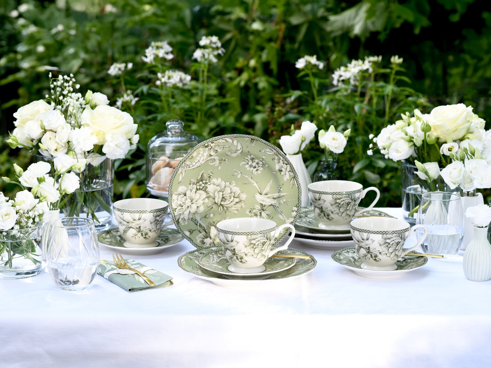Elegantes Kaffee- und Tafelservice in Grün mit floralem Muster, Tassen und Teller auf weiß gedecktem Tisch mit weißen Blumen und Gläsern im Garten, frontal aus leicht erhöhter Perspektive