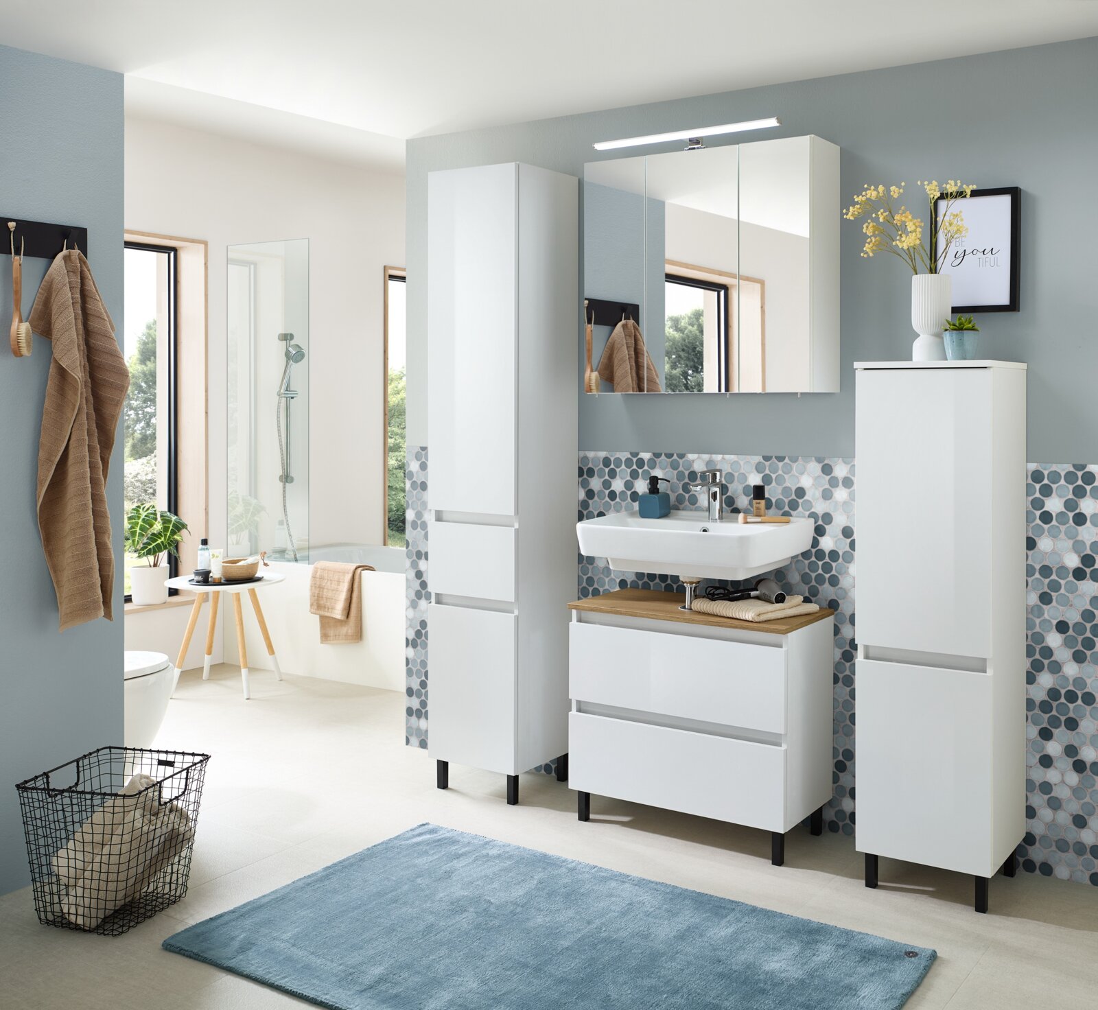 Modernes Badmöbel-Set in Weiß mit Waschtisch, Unterschrank mit Schubladen, Spiegelschrank mit LED-Leuchte sowie zwei Hochschränken auf schwarzen Füßen, fotografiert in leichter Frontansicht aus Augenhöhe.