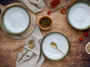 Drei Keramikschalen in Weiß mit grünem Rand auf rustikalem Holztisch, dekoriert mit Oliven, Kirschtomaten, Holzlöffel und goldenem Besteck, fotografiert aus der Vogelperspektive (Top-Down).