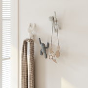 Drei Wandhaken in Form von kletternden Figuren, an einer hellen Wand montiert. Einer der Haken hält ein kariertes Handtuch, während ein anderer eine hängende Dekoration trägt. Perspektive von vorne.