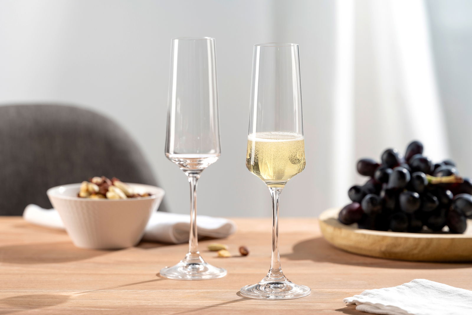 Leonardo Set van 6 champagneglazen PALADINO Zwei elegante Sektgläser auf einem Tisch, eines gefüllt mit Sekt, im Hintergrund eine Schale mit Nüssen und ein Teller mit Trauben, seitliche Perspektive.