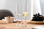 Zwei elegante Sektgläser auf einem Tisch, eines gefüllt mit Sekt, im Hintergrund eine Schale mit Nüssen und ein Teller mit Trauben, seitliche Perspektive.