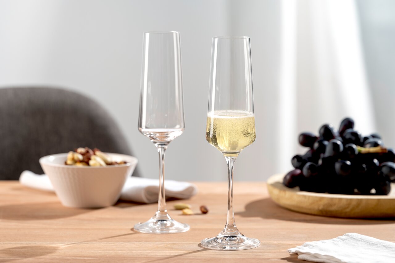 Twee elegante champagneglazen op een tafel, waarvan één gevuld met champagne, op de achtergrond een schaal met noten en een bord met druiven, zijaanzicht.