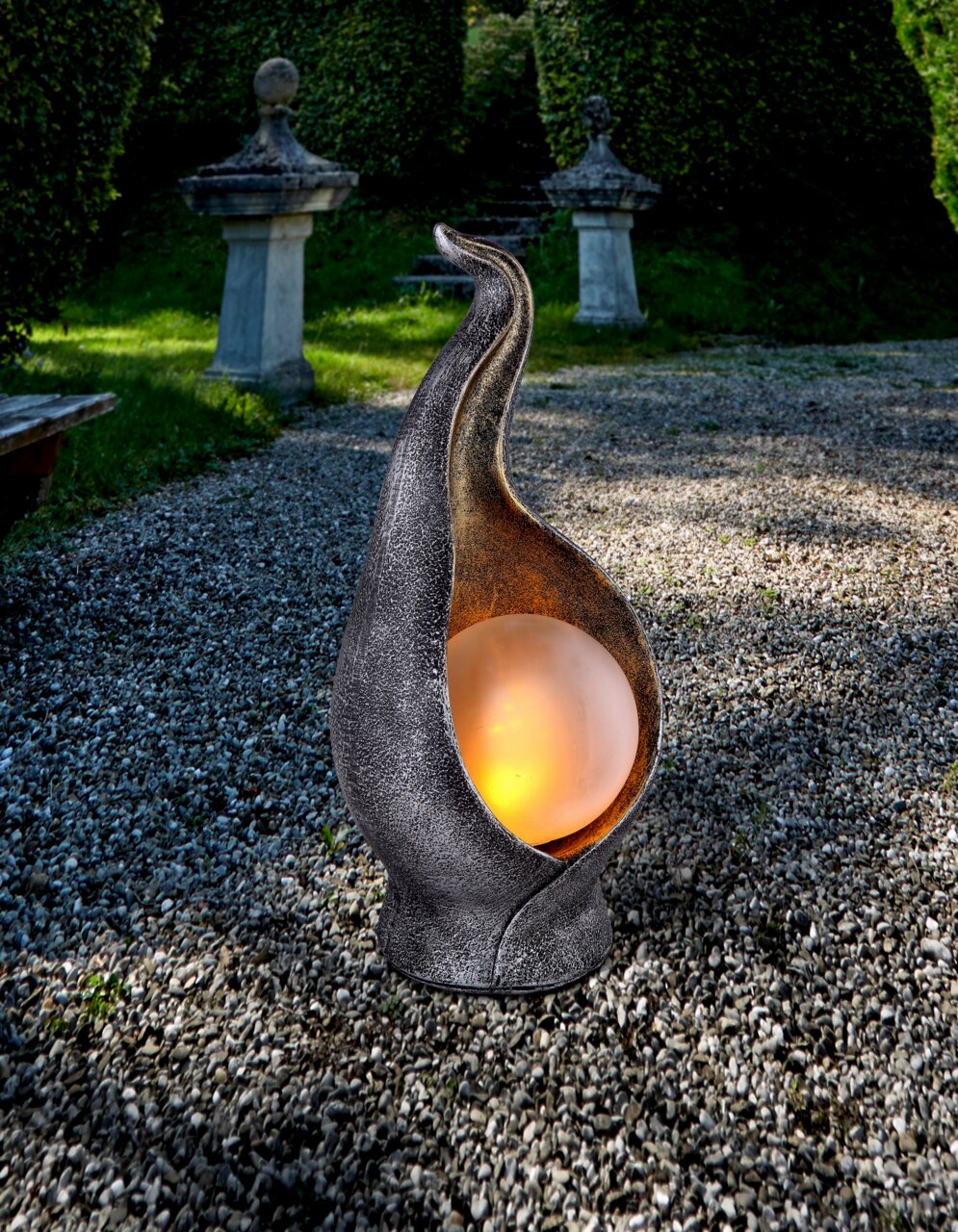Solarleuchte Oliver in einem Garten, aus der Perspektive von vorne, mit einer leuchtenden Kugel in einer kunstvoll geschwungenen Metallhülle.
