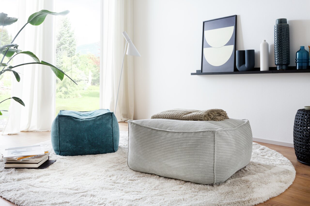 Zwei quadratische Hocker in einem modernen Wohnzimmer, einer in Blau und einer in Grau, auf einem runden Teppich vor einem großen Fenster. Perspektive von schräg oben.