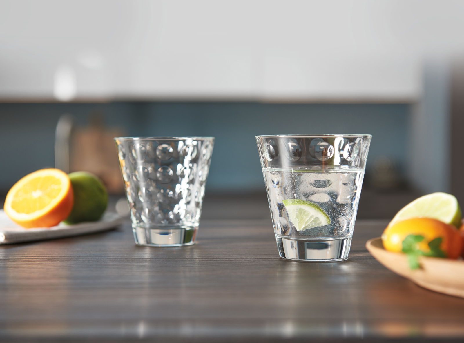 Leonardo Whiskybecher OPTIC Zwei Trinkgläser Optic 215ml auf einem Tisch, eines gefüllt mit Wasser und Limettenscheibe, im Hintergrund unscharf Obst und Küche, seitliche Perspektive.
