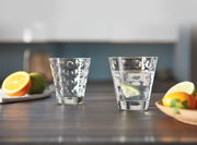 Zwei Trinkgläser Optic 215ml auf einem Tisch, eines gefüllt mit Wasser und Limettenscheibe, im Hintergrund unscharf Obst und Küche, seitliche Perspektive.